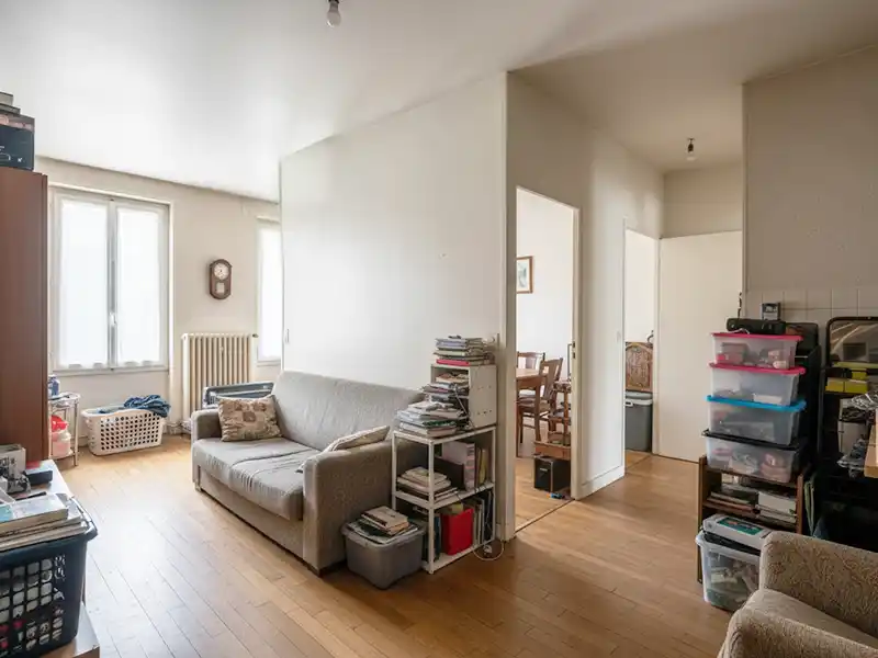 Pièce mal organisée Un salon lumineux mais mal organisé avec des planchers en bois clair, un canapé gris, des livres et des bacs de rangement sur des étagères, et une porte ouverte sur la salle à manger. La lumière du soleil passe à travers deux fenêtres, mettant en valeur les paniers à linge dans l'espace.