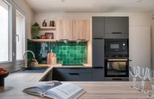 Cuisine moderne après une rénovation complète de la cuisine, avec un dosseret en carrelage vert, des armoires en bois clair et foncé, un four et un micro-ondes encastrés, un évier près d'une fenêtre et un livre de cuisine ouvert à côté de verres à vin vides sur le comptoir.