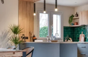 Cuisine moderne avec dosseret en carreaux verts, armoires en bois, îlot central avec verres et assiettes, lampes suspendues au plafond, plante dans le coin, et une fenêtre qui remplit cette rénovation cuisine américaine de lumière naturelle.