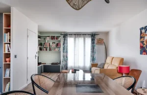 Salon moderne après une rénovation pièce intérieure, comprenant un canapé beige, un lampadaire et une lampe de table rouge. Des bibliothèques bordent le mur, une table à manger avec des chaises tressées au premier plan, et des rideaux à motifs encadrent de grandes fenêtres qui laissent entrer la lumière naturelle.