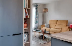 Un salon confortable après une rénovation, avec un canapé jaune, des coussins à motifs, une table basse en verre, un lampadaire et un petit chien couché sur le tapis. Des étagères et un meuble de cuisine bleu sont partiellement visibles au premier plan.