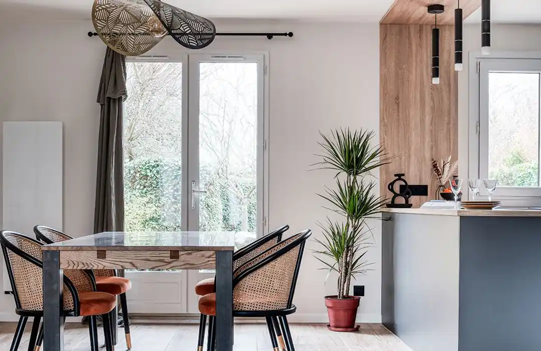 Un coin repas moderne, con&ccedil;u dans le cadre d'un s&eacute;jour de r&eacute;novation r&eacute;cente, comprend une table en verre et quatre chaises tress&eacute;es. De grandes fen&ecirc;tres inondent l'espace de lumi&egrave;re, tandis qu'une plante en pot et des lampes suspendues lui conf&egrave;rent un style &eacute;l&eacute;gant.