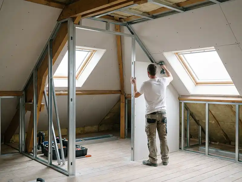 Travaux aménagement des combles - Destpec Dans le cadre de travaux d'aménagement des combles, une personne en tenue de travail pose des panneaux de placoplâtre sur une ossature métallique dans une pièce mansardée lumineuse, avec des plafonds en angle, deux grandes fenêtres à vasistas et des outils de construction sur le sol en bois.