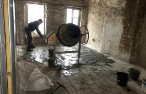 Une personne effectue des travaux de maçonnerie, étalant du béton humide sur un sol carrelé à l'intérieur d'une pièce inachevée aux murs en pierres apparentes. À proximité, une bétonnière et des seaux sont posés, tandis que la lumière du jour pénètre par deux fenêtres.